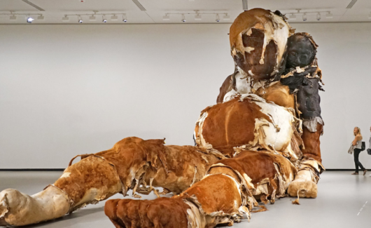 De sculptuur op de afbeelding is "Giant No. 3" van de Chinese kunstenaar Zhang Huan. Het monumentale werk toont een zittende figuur, lijkend op een zwangere bedelaar of een sjamaan, met een kleiner figuur, een zelfportret van de kunstenaar, bij haar nek.